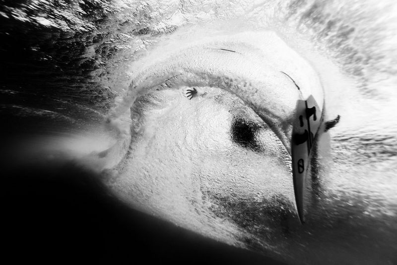A black and white photo taken from under the water by Todd Glasser, showing a surfer with just his board and his hand making contact with the water.