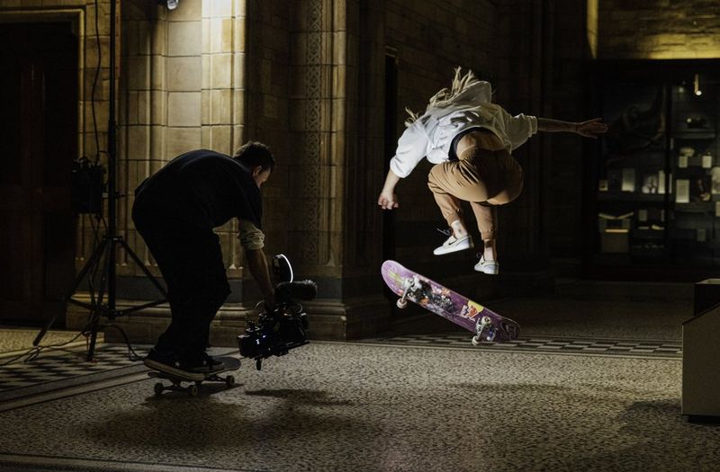 Un uomo su uno skateboard regge una videocamera per registrare una skater che esegue trick sul suo skateboard.