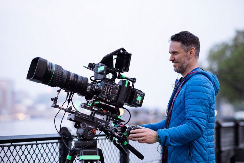 A cinematographer in a blue puffer coat operating a Canon EOS C300 Mark III camera with a CN10X25 IAS S lens next to a metal railing on the River Thames, filming the London wingsuit event.
