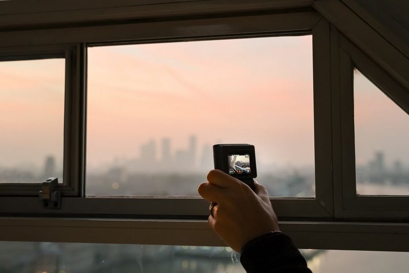 A user's hand holds a Canon PowerShot V10 vlogging camera and points through a window at the river and London skyline behind, ready to film the London wingsuit event.