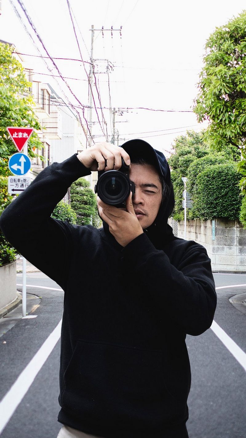 A man in a black top with the hood up, holds a Canon camera up as though taking a photograph. Behind him is a street scene with Japanese road signs.