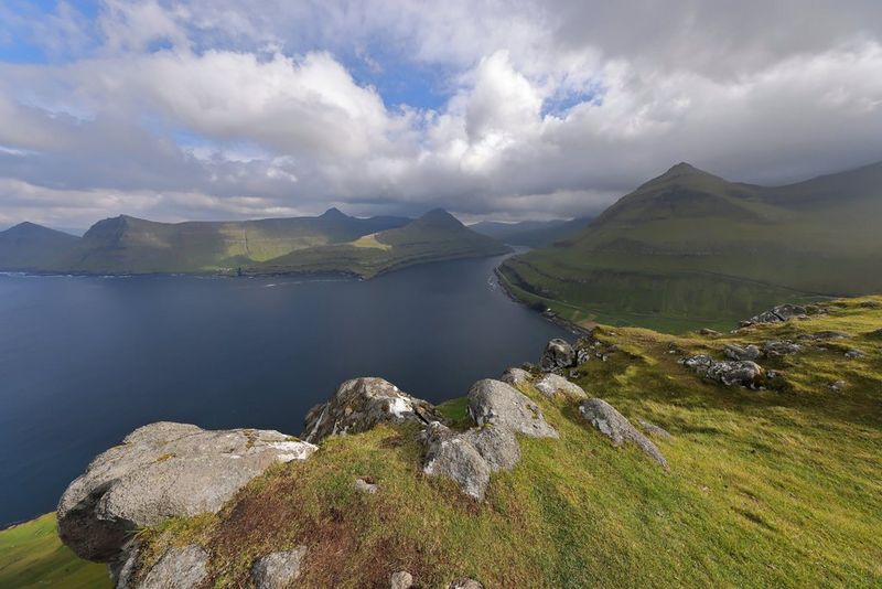 A cloudy sky over a lake, surrounded by bright green hills and mountain peaks.