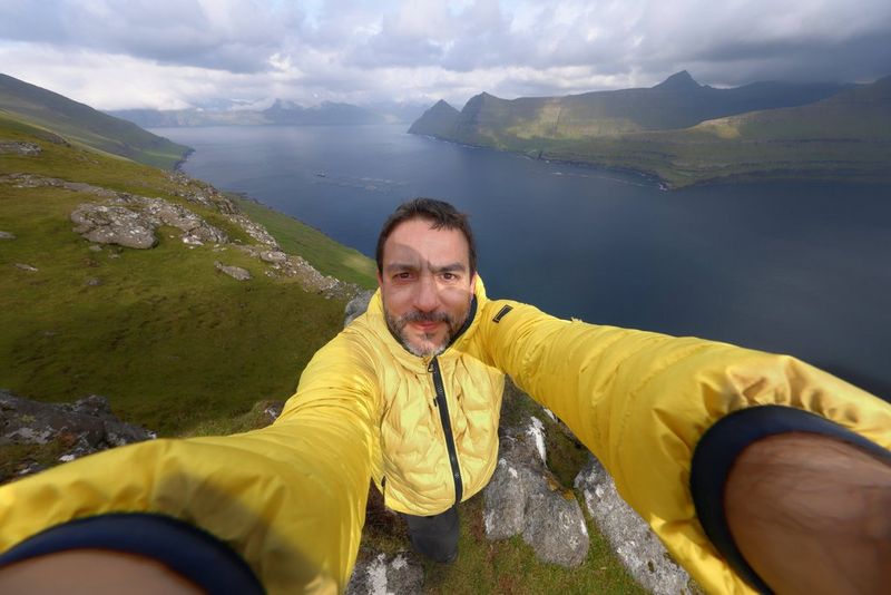 Joel Santos takes a selfie with a Canon camera attached with a Canon RF 10-20mm F4L IS STM lens, a lake and hills visible behind him. 