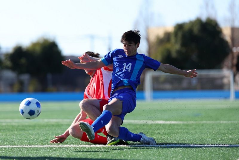 Een actiefoto gemaakt met een Canon RF 100-300MM F2.8L IS USM van twee voetballers, een in het blauw en een in het rood, die tegen elkaar botsen wanneer de eerste de bal wegtrapt.