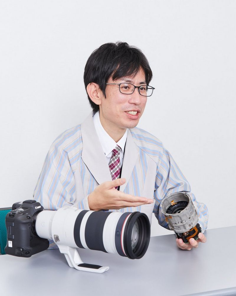 Een ontwikkelaar houdt een deel van het interne mechanisme van de RF 100-300MM F2.8L IS USM vast, terwijl het complete objectief voor hem op tafel ligt, bevestigd aan een camera.