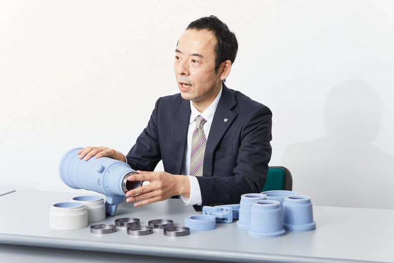 Een man houdt een prototype van de RF 100-300MM F2.8L IS USM vast, met verschillende prototypes van onderdelen op de tafel voor hem.