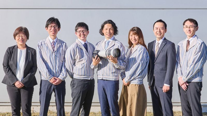 Zeven mensen, de ontwikkelaars van de Canon RF 100-300MM F2.8L IS USM, staan naast elkaar en de man in het midden houdt een camera vast met het objectief