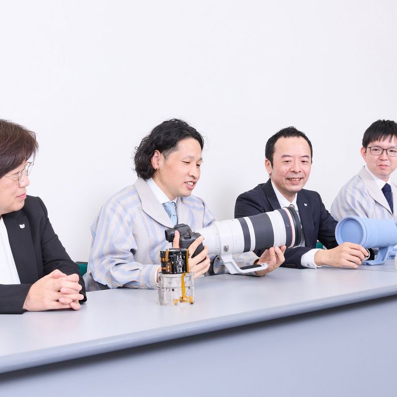 Four of the developers of the Canon RF 100-300mm F2.8L IS USM sit at a table, one of them holding a camera with the lens attached.
