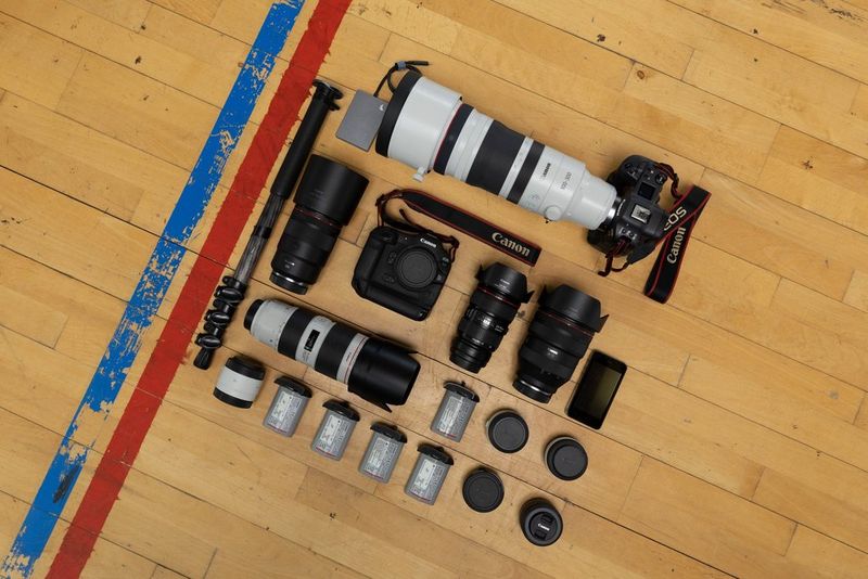 A selection of Canon cameras and lenses plus other camera equipment are laid out in a square pattern on a gymnasium floor, next to two stripes in red and blue marked on the floor.