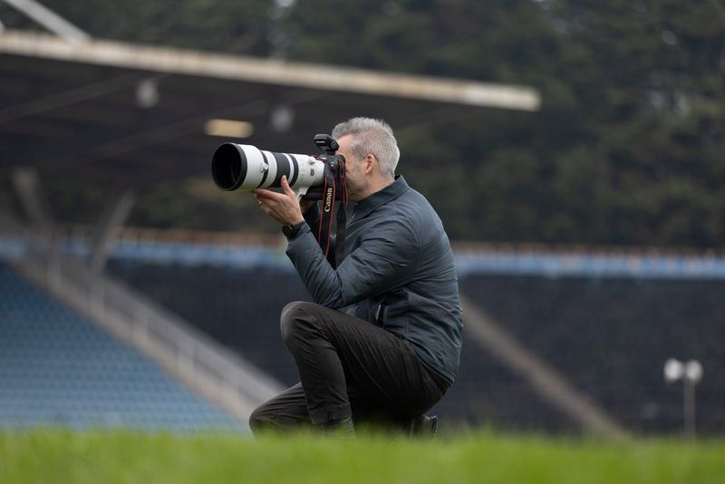 Photographer Julian Finney crouches on grass in the middle of an athletics stadium, holding up a Canon EOS R3 with a Canon RF 100-300mm F2.8L IS USM lens to his face.