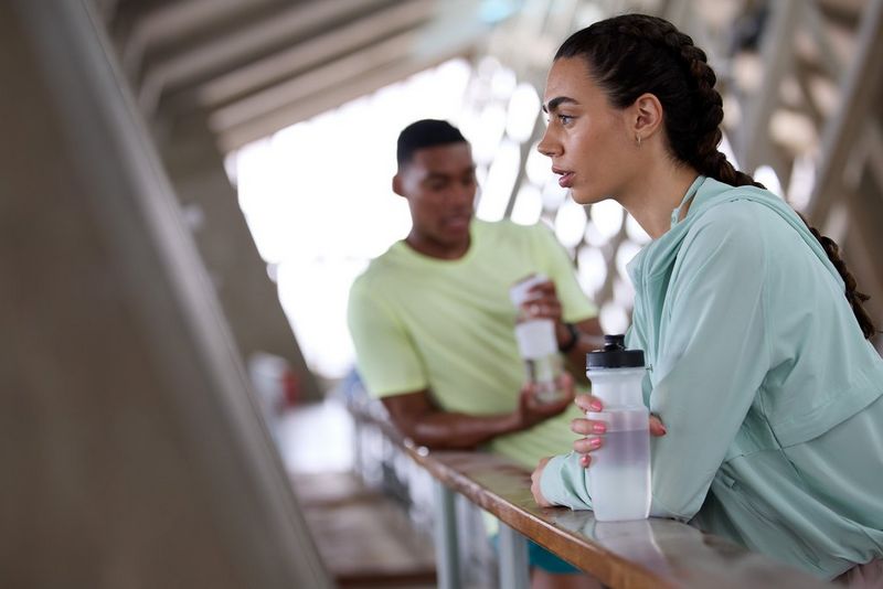A woman in a tracksuit leans against a wooden rail with a water bottle in her hand. She is in sharp focus, while the figure in the background is blurred