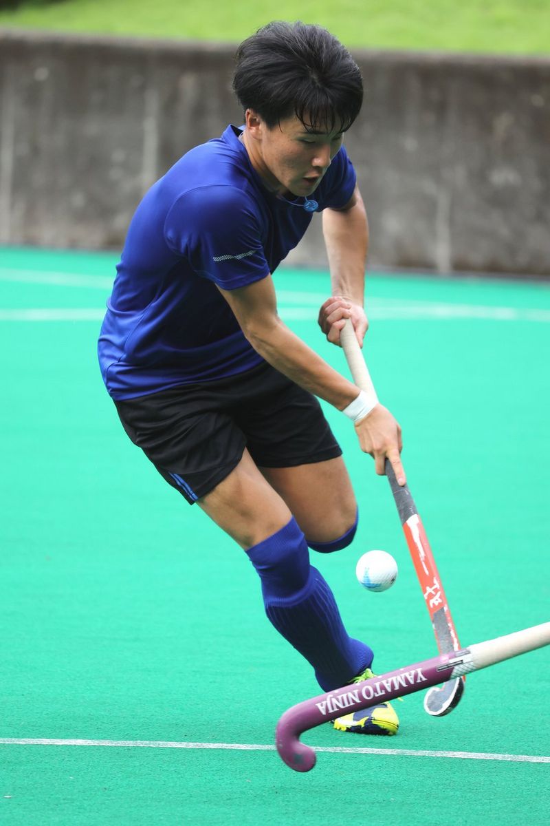Een hockeyspeler in een blauw shirt en hoge blauwe sokken strijdt om de bal op een kunstveld.