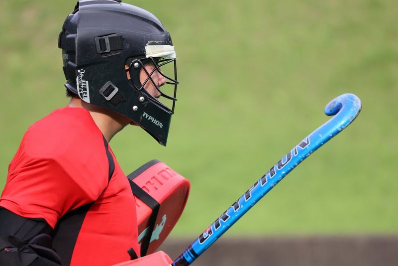 Zijaanzicht van een hockeyspeler in een rood shirt en een zwarte helm, die een blauwe hockeystick omhoog houdt.