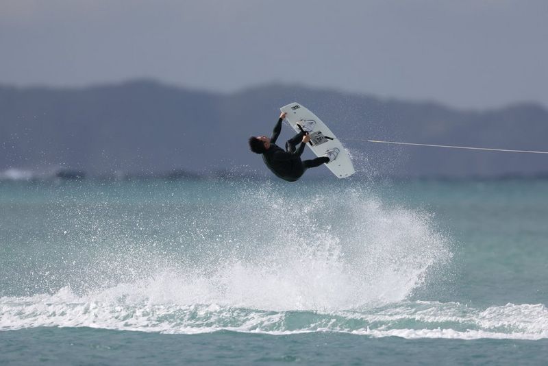 A wakeboarder leaps into a trick above the ocean as white spray crashes below him. Action sports photo taken with a Canon RF 1200mm F8L IS USM lens.