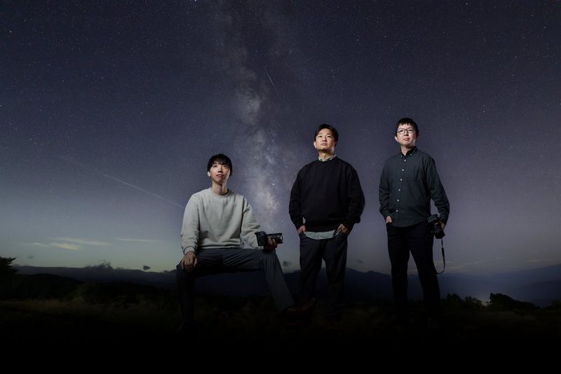 Les trois ingénieurs responsables de la conception de l'objectif ultra grand-angle RF 14mm F1.4L VCM de Canon, devant une image du ciel étoilé.