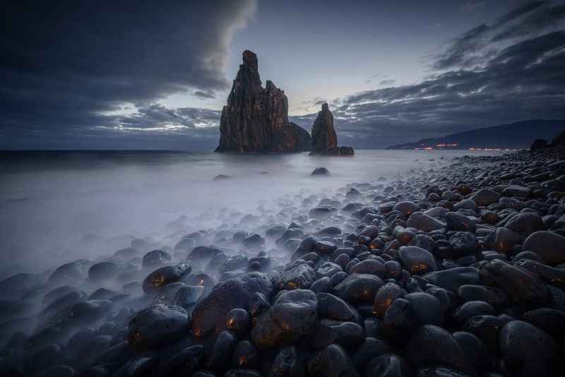 A low-light seascape showing blurred waves flowing around rounded shoreline rocks, with illuminated buildings in the distance. Taken on a Canon EOS R5 Mark II.