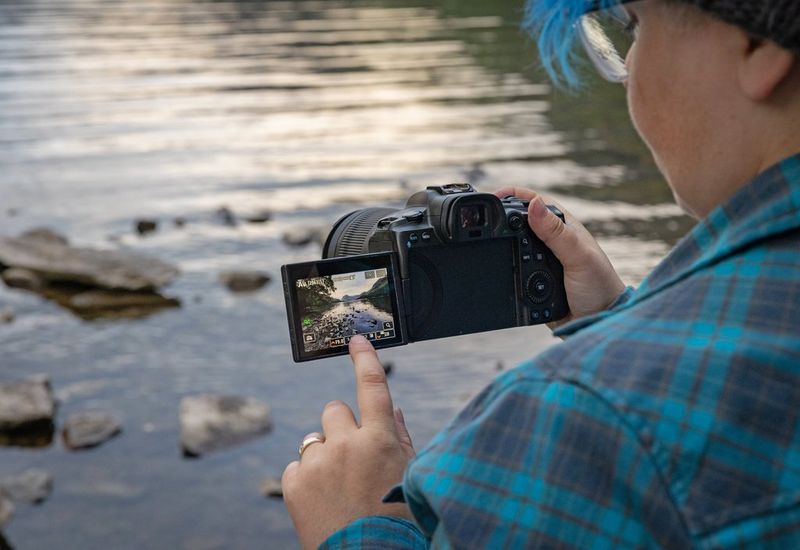 Una vista da sopra la spalla di un fotografo mentre tocca lo schermo LCD di una fotocamera Canon con un obiettivo Canon RF 15-30mm F4.5-6.3 IS STM. Sullo schermo è visibile una scena con un torrente roccioso.