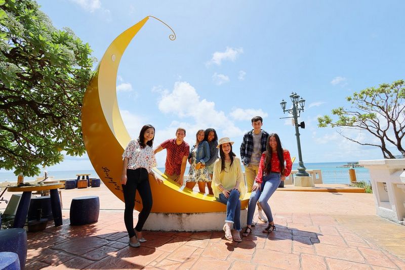 Групна фотографија од луѓе собрани околу голема жолта скулптура во форма на банана на сончев поплочен плоштад со видливо море во заднина. Фотографирано со широкоаголен објектив Canon RF 16mm F2.8 STM.