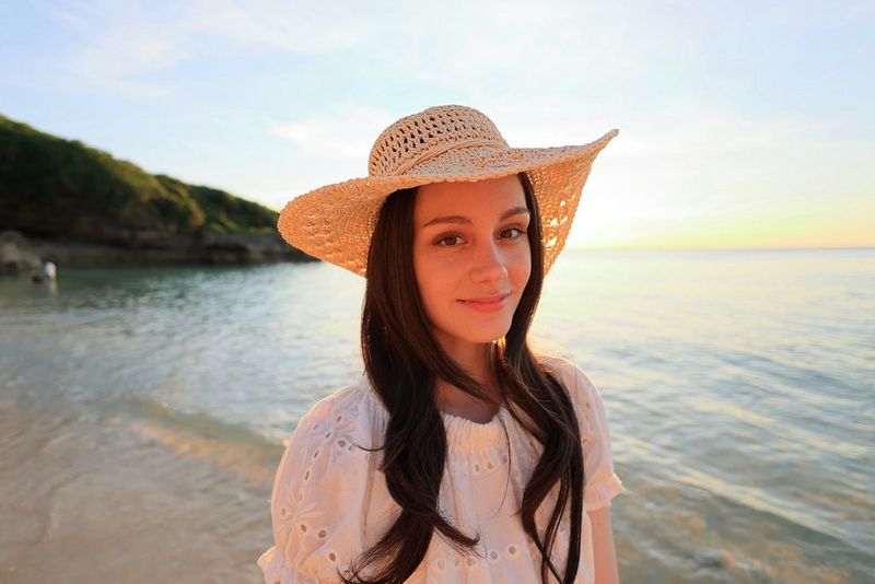דיוקן של אישה עומדת בחוף הים וחובשת כובע שמש, שצולם בעדשת Canon RF 16mm F2.8 STM.