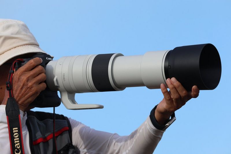 A photographer in a sunhat holds a Canon camera with a white-coated Canon RF 200-800mm F6.3-9 IS USM lens attached.