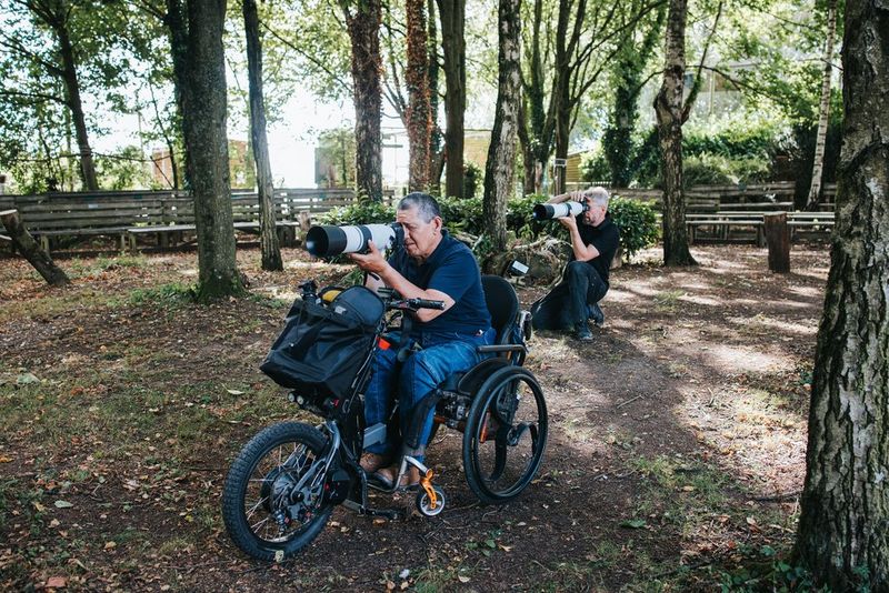 In a clearing surrounded by trees, two photographers, one in a wheelchair, use their Canon cameras with the RF 200-800mm F6.3-9 IS USM lens attached. 