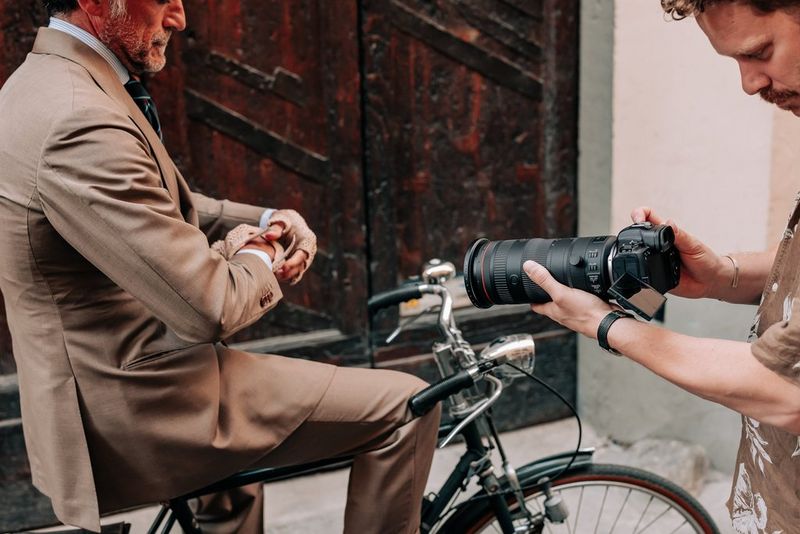 Hands holding a Canon EOS R5, with a Canon RF 24-105mm F2.8L IS USM Z lens attached, to film a besuited man on a bicycle.