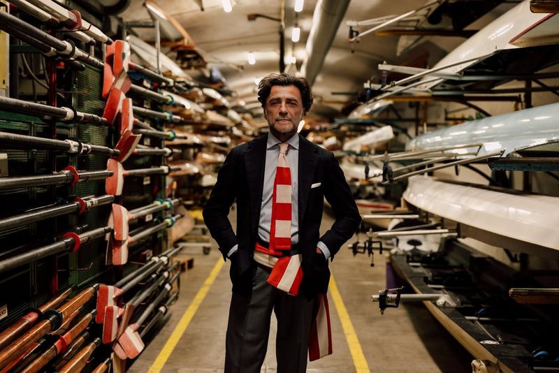 A man stands in a room with his hands in his pockets, looking directly into the camera lens, surrounded by rowing boats and equipment.