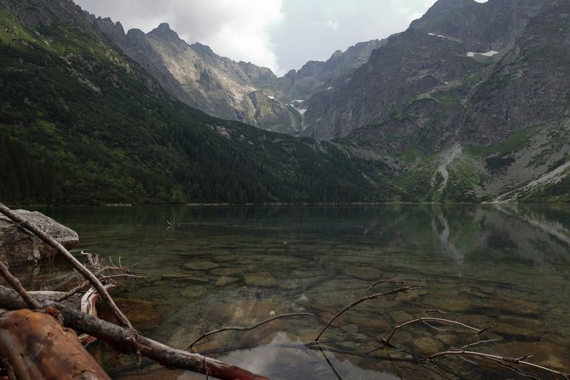 Чистое мелководное озеро, ветви на переднем плане и горные вершины на фоне.