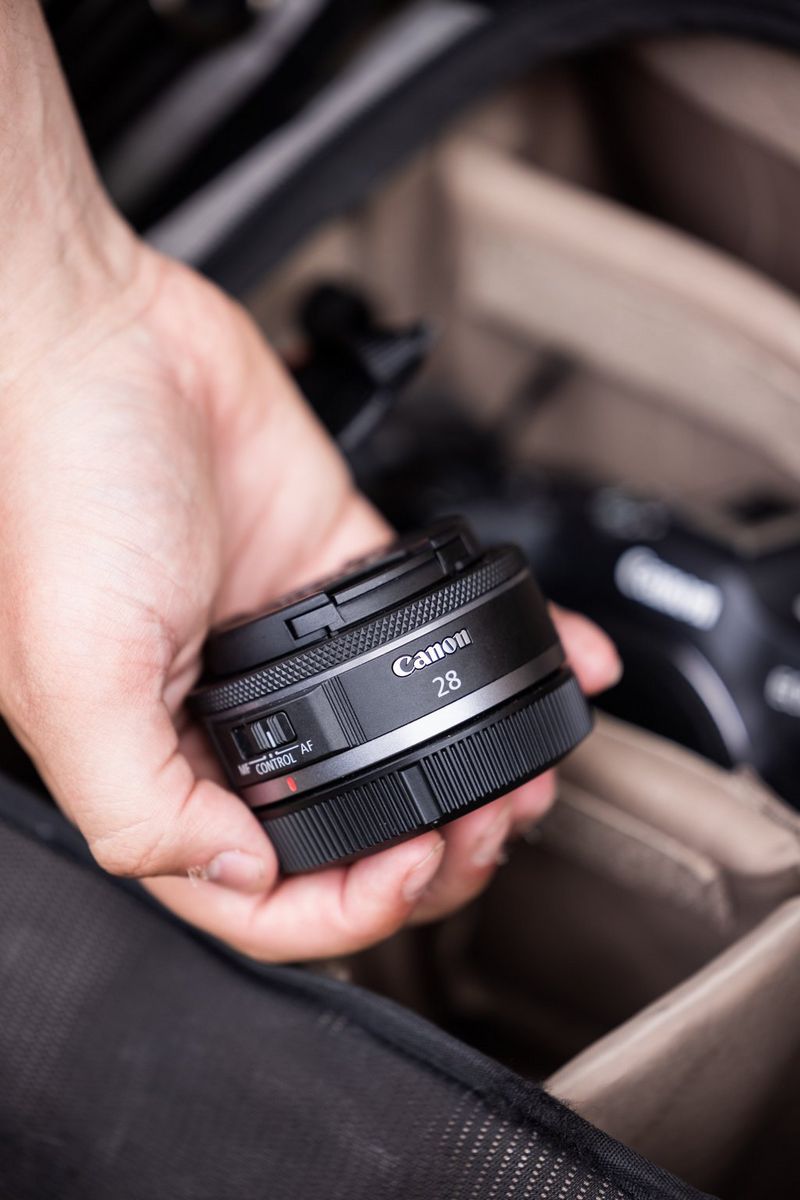 A user's hand holds a Canon RF 28mm F2.8 STM prime lens over a camera kit bag, which is slightly out of focus.