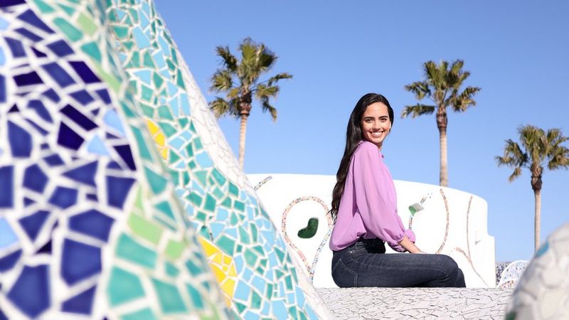 דיוקן של אישה מחייכת שיושבת ליד פסיפס אריחים בצבעי כחול ואקווה וברקע שמיים כחולים וצלולים ועצי דקל. צולם בעדשת Canon RF 28mm F2.8 STM.