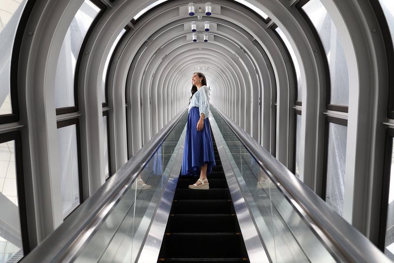 A woman standing on a covered escalator in a blue skirt, taken on an EOS R8.