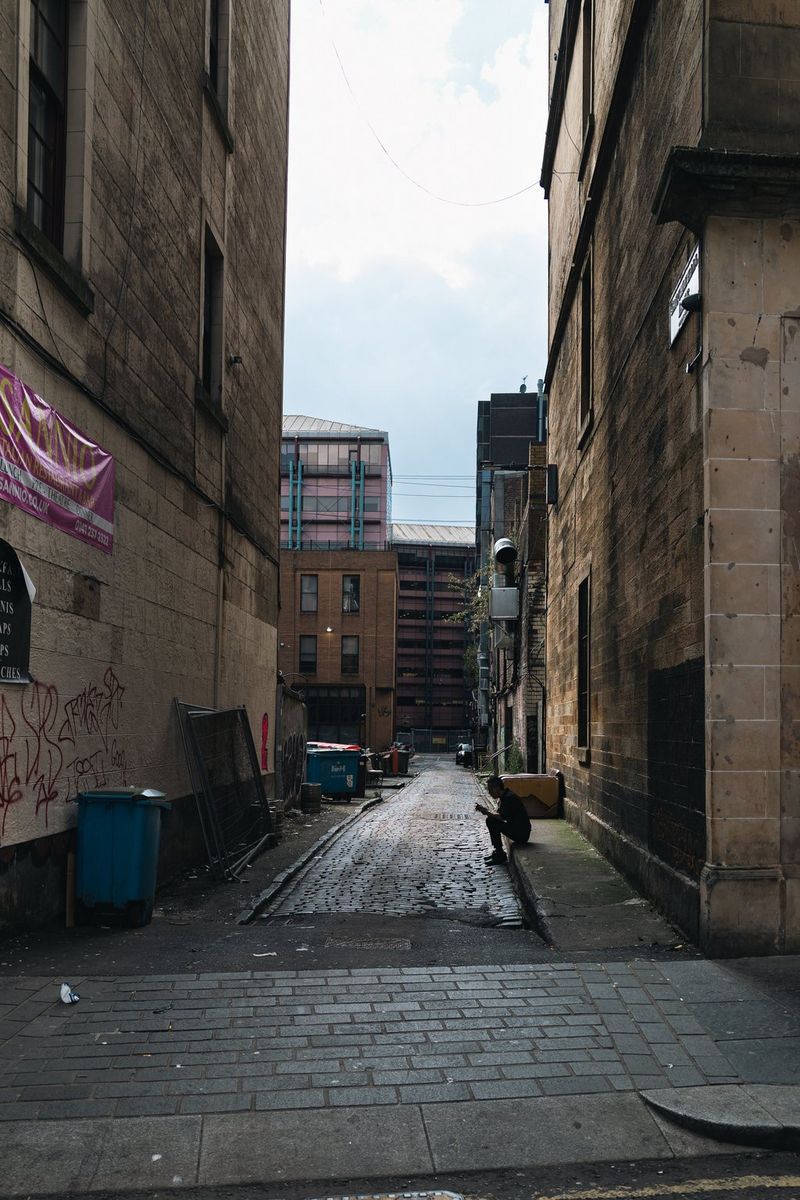 Человек сидит на бордюре в тени между двух высотных зданий в солнечный день и смотрит в телефон. Снято Андресом Макнилом с помощью объектива Canon RF 28mm F2.8 STM.