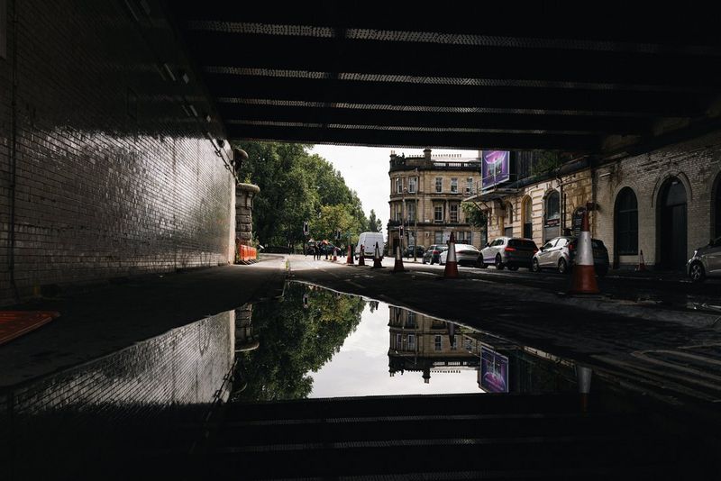 Shot by street photographer Andres McNeill from underneath a bridge, a building and trees are reflected in a large puddle of water in the road.