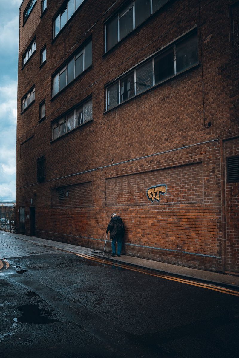 Street photographer Andres McNeill captures a street photography image of an elderly man walking along the pavement with a stick, dwarfed by the red brick walls of a high-rise building.