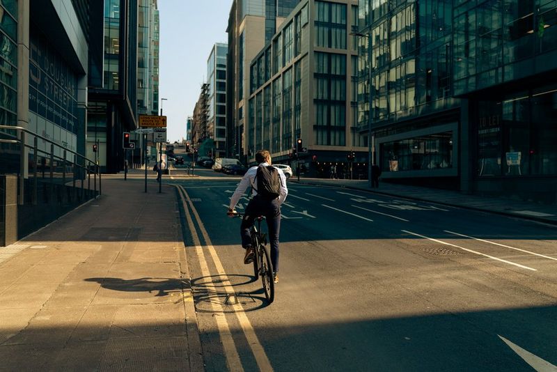 A man riding a bike down a deserted city street surrounded by modern glass buildings during the daytime, with sunlight casting long shadows across the road.