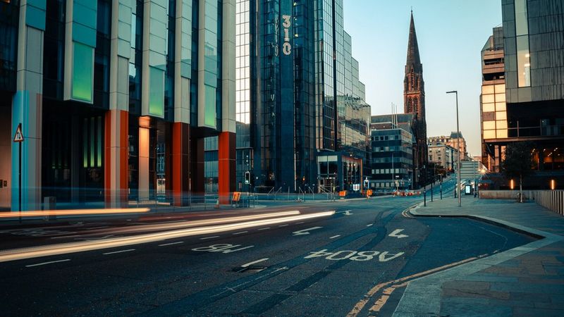 Следы от автомобильных фар, запечатленные ранним вечером на улице с высотными зданиями; фото создано Андресом Макнилом с помощью объектива Canon RF 28mm F2.8 STM.