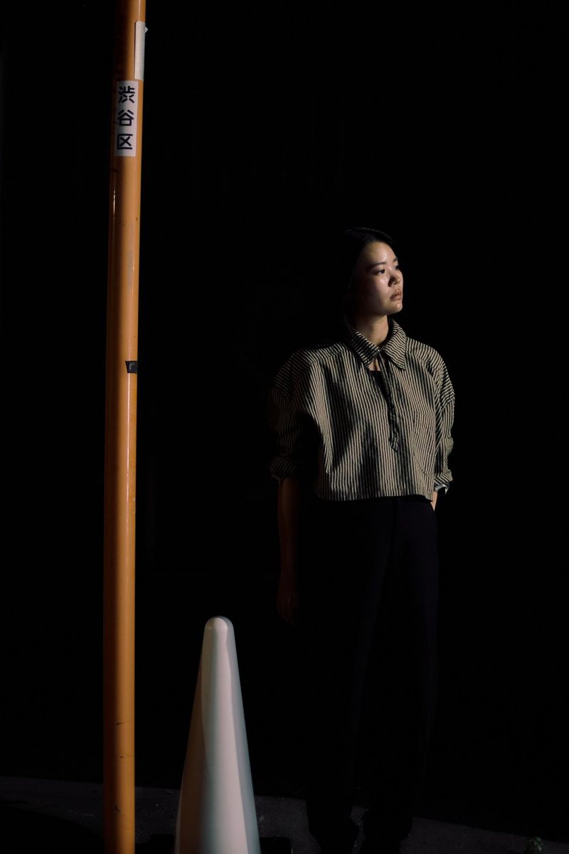 Портрет жінки в темних штанях і блузці з темними й світлими вертикальними смугами, зроблений за слабкого освітлення за допомогою об’єктива з фіксованою фокусною відстанню Canon RF 45mm F1.2 STM.