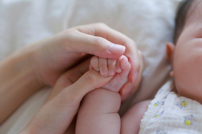 A close-up of a baby's hand gripping an adult's thumb, with a second adult's hand enfolding both, taken with a Canon RF 45mm F1.2 STM prime lens.