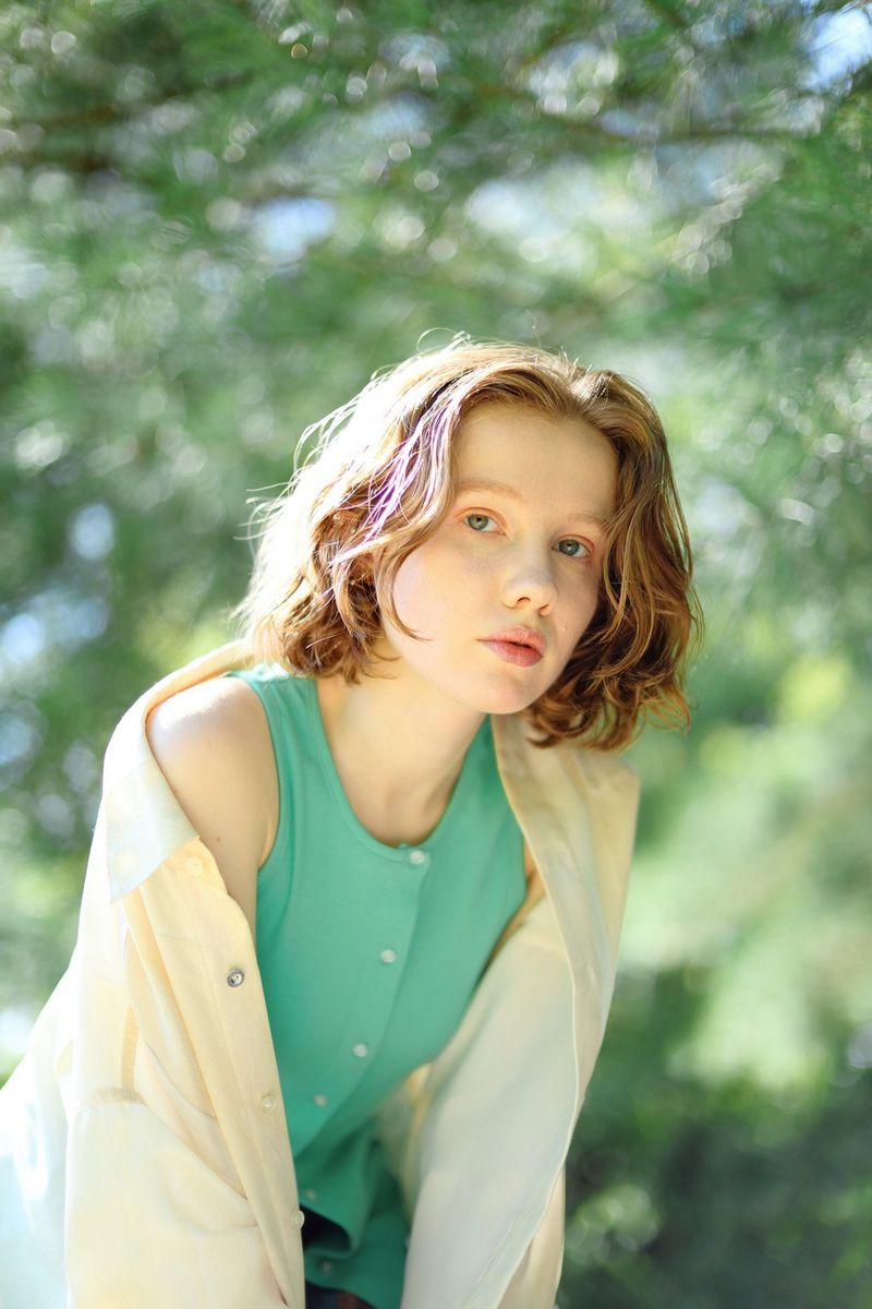 A three-quarter-length portrait of a young woman leaning in towards the camera on a sunlit day against a diffuse backdrop of trees, taken with a Canon RF 45mm F1.2 STM lens.