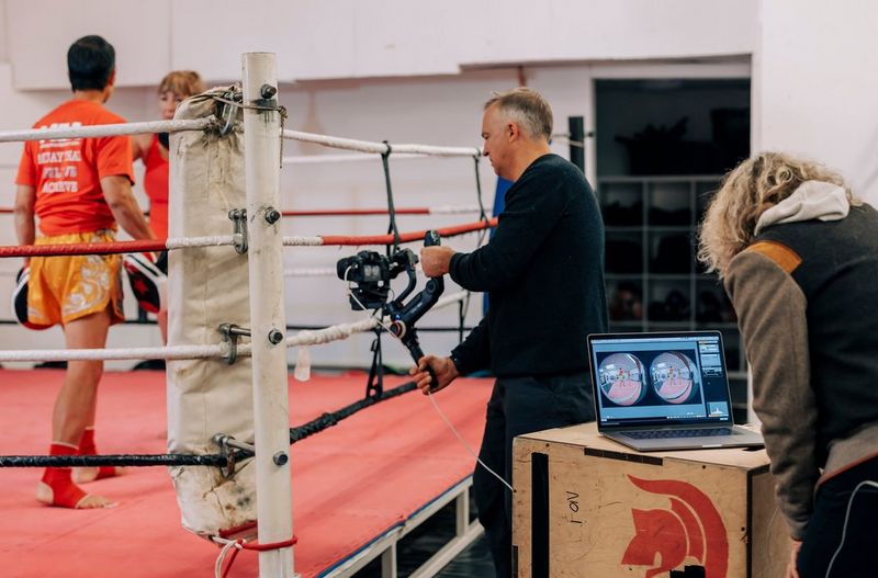 A man using a Canon EOS R5 camera with Canon RF 5.2mm F2.8L Dual Fisheye lens films two people in a boxing ring. Two circular fisheye images are visible on a laptop screen in the foreground.