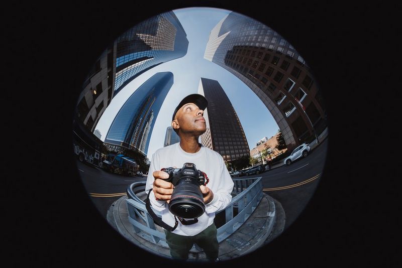  A circular fisheye image of photographer Atiba Jefferson holding a Canon EOS R5 Mark II and RF 7-14mm F2.8-3.5L FISHEYE STM, the buildings in the background distorted.