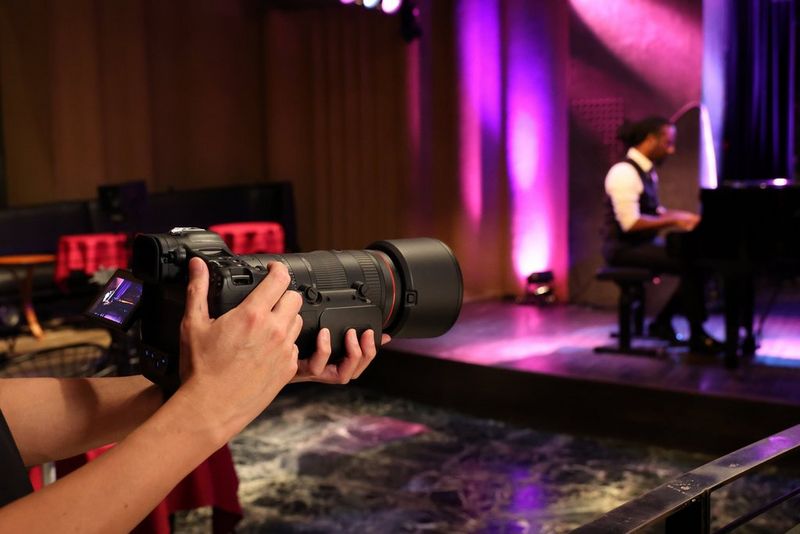 A user’s hands hold a Canon camera with an RF-70-200mm-F2-8L-IS-USM-Z lens with Power Zoom filming a person playing piano on a stage in the background.