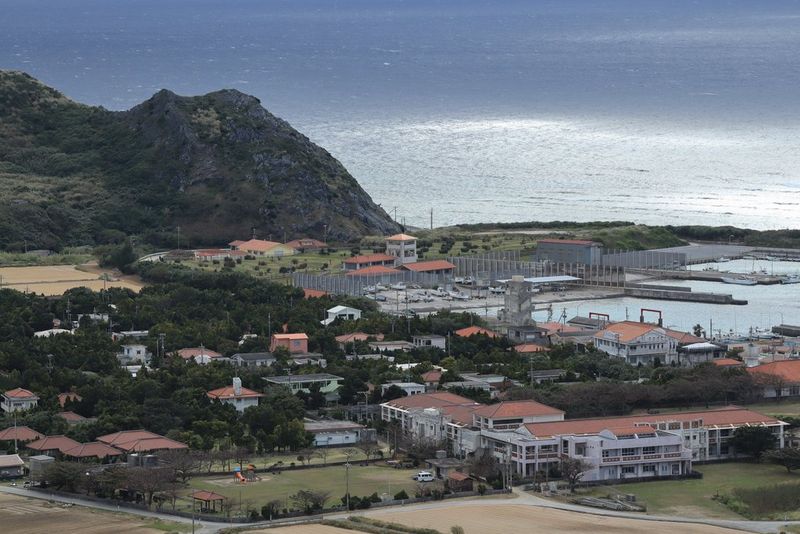Imagen tomada a una longitud focal de 100 mm que muestra unos edificios junto a una ensenada en la costa, con una colina cubierta de hierba al fondo.