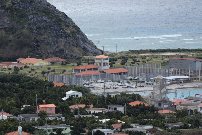 Imagen tomada a una longitud focal de 200 mm que muestra un astillero rodeado por una valla alta y otros edificios junto a una ensenada en la costa, con una colina cubierta de hierba elevándose detrás.