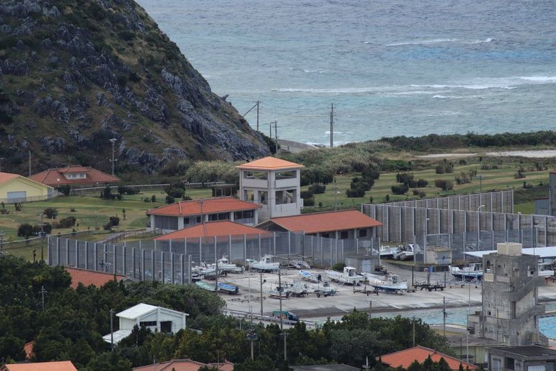Imagen tomada a una longitud focal de 300 mm que muestra un astillero rodeado por una valla alta, con otros edificios en primer plano y una colina elevándose detrás, y el mar al fondo.