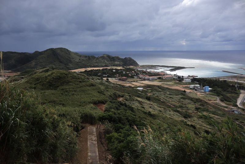 Imagen tomada a una longitud focal de 18 mm que muestra colinas cubiertas de hierba que descienden hacia una ensenada en la costa, con algunos edificios en la distancia.
