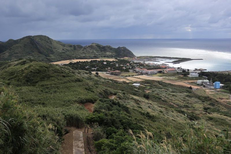 Imagen tomada a una longitud focal de 24 mm que muestra colinas cubiertas de hierba que descienden hacia una ensenada en la costa, con algunos edificios en la distancia.