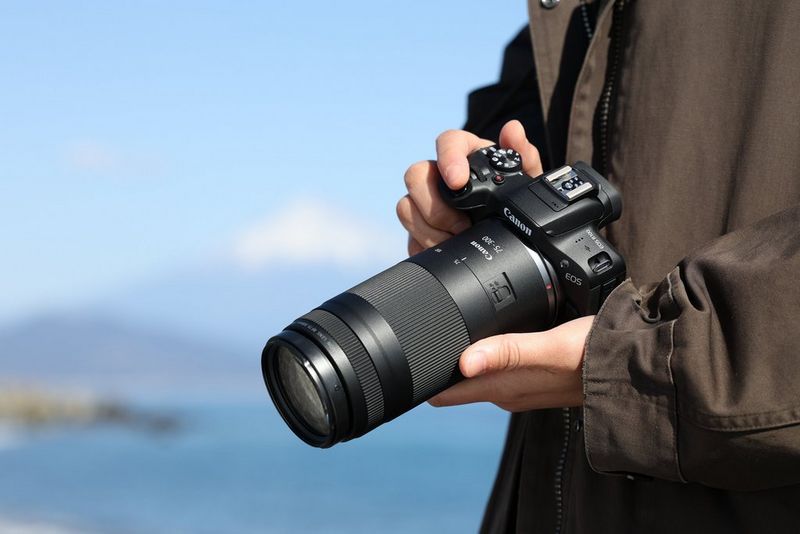 Un utente tiene in mano una fotocamera Canon con obiettivo Canon RF 75-300mm F4.0-5.6. Una vetta innevata si staglia sopra uno specchio d'acqua sullo sfondo.