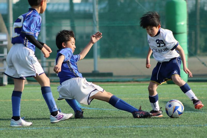 Foto de acción de un joven futbolista estirándose para alcanzar el balón con un pie, casi horizontal al suelo, entre otros jugadores. Imagen tomada con un teleobjetivo Canon RF 75–300mm F4–5.6 a una longitud focal de 230 mm.