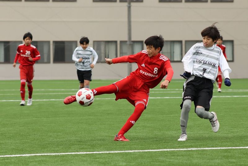 Photo d'action d'un jeune footballeur en tenue rouge sur le point de frapper le ballon à hauteur de cuisse, avec d'autres joueurs également visibles sur le terrain, prise avec un objectif zoom Canon RF 75-300mm F4-5.6.